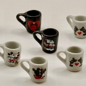 May include: A collection of nine miniature ceramic mugs in black and white. Each mug features a small handle and a decorative graphic of cartoon mice characters, some with red heart accents, arranged in a scattered pattern on a plain surface.