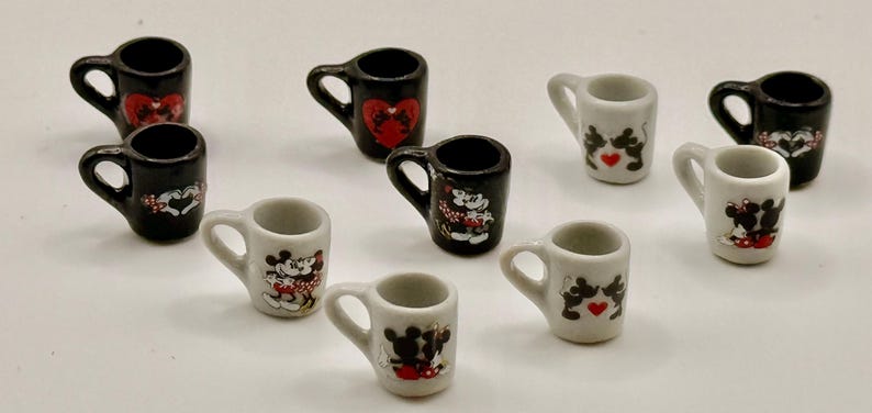 May include: A collection of nine miniature ceramic mugs in black and white. Each mug features a small handle and a decorative graphic of cartoon mice characters, some with red heart accents, arranged in a scattered pattern on a plain surface.