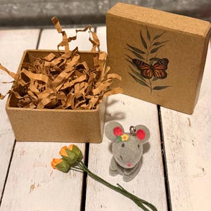 May include: A grey mouse ornament with red ears and a floral crown sits beside a small gift box featuring a butterfly illustration. A small box filled with brown paper shreds and a bunch of orange and green flowers are also present.