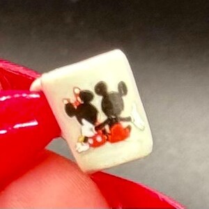 May include: A miniature white mug featuring a graphic illustration of two cartoon characters, one with black ears and red shorts and another with a red bow, sitting side by side. The small mug has a cylindrical shape and a smooth, off-white finish.