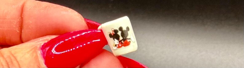May include: A miniature white mug featuring a graphic illustration of two cartoon characters, one with black ears and red shorts and another with a red bow, sitting side by side. The small mug has a cylindrical shape and a smooth, off-white finish.