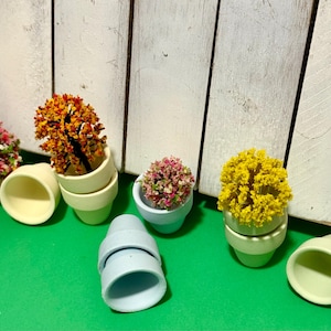 May include: Miniature ceramic flower pots in various colors, including light blue, yellow, and pale green, filled with colorful artificial flowers. The pots are arranged on a green surface against a white wooden backdrop.