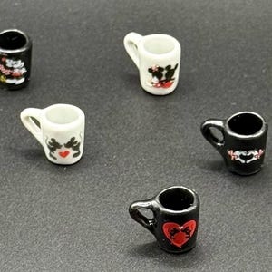 May include: A collection of miniature ceramic mugs in black and white, each featuring small graphic prints of cartoon characters and red heart motifs. The mugs are arranged in a scattered pattern, showcasing various designs on their exterior surfaces.