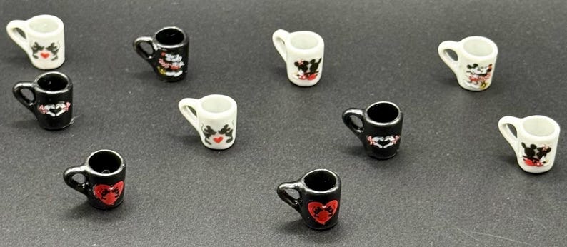 May include: A collection of miniature ceramic mugs in black and white, each featuring small graphic prints of cartoon characters and red heart motifs. The mugs are arranged in a scattered pattern, showcasing various designs on their exterior surfaces.