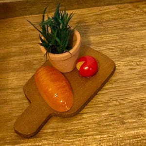Puede incluir: Tabla de cortar de madera en miniatura con un pan, una rueda de queso rojo y una pequeña planta en maceta. La planta tiene hojas verdes y está en una maceta de terracota. El pan es naranja y brillante.