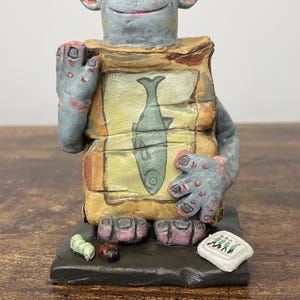 Puede incluir: Una figura caprichosa pintada a mano de una criatura parecida a un trol. La figura tiene piel gris, manos y pies grandes, y sostiene una bolsa con una ilustraci&oacute;n de un pez. Peque&ntilde;os insectos pintados y un letrero en la base.