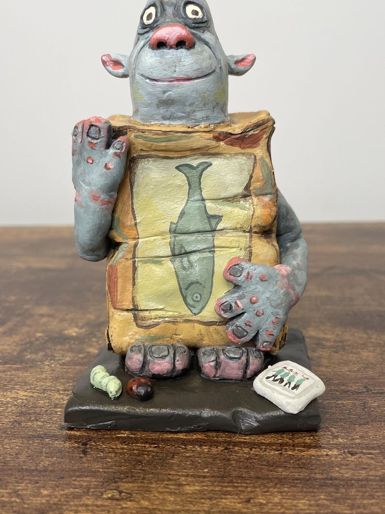Puede incluir: Una figura caprichosa pintada a mano de una criatura parecida a un trol. La figura tiene piel gris, manos y pies grandes, y sostiene una bolsa con una ilustraci&oacute;n de un pez. Peque&ntilde;os insectos pintados y un letrero en la base.