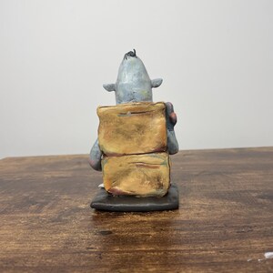 Puede incluir: Escultura de cer&aacute;mica hecha a mano de una criatura con cabeza gris, orejas peque&ntilde;as y un mech&oacute;n de pelo negro. La criatura est&aacute; unida a dos formas rectangulares apiladas de color marr&oacute;n dorado. La escultura est&aacute; sobre una base negra.