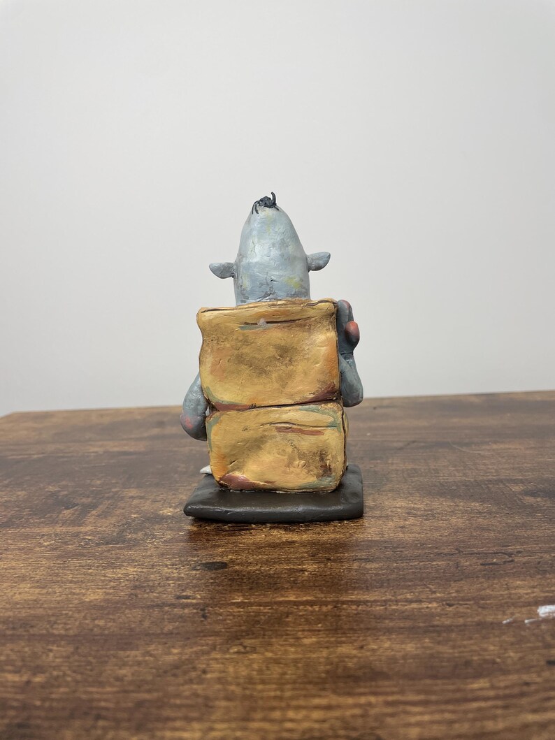 Puede incluir: Escultura de cer&aacute;mica hecha a mano de una criatura con cabeza gris, orejas peque&ntilde;as y un mech&oacute;n de pelo negro. La criatura est&aacute; unida a dos formas rectangulares apiladas de color marr&oacute;n dorado. La escultura est&aacute; sobre una base negra.