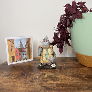 Puede incluir: Una figura caprichosa de una criatura sosteniendo un paquete con una ilustraci&oacute;n de pescado, junto a una tarjeta de felicitaci&oacute;n con un paisaje urbano europeo. La escena est&aacute; ambientada sobre una superficie de madera, con una planta decorativa en una maceta verde y marr&oacute;n.