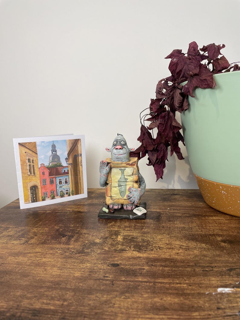 Puede incluir: Una figura caprichosa de una criatura sosteniendo un paquete con una ilustraci&oacute;n de pescado, junto a una tarjeta de felicitaci&oacute;n con un paisaje urbano europeo. La escena est&aacute; ambientada sobre una superficie de madera, con una planta decorativa en una maceta verde y marr&oacute;n.
