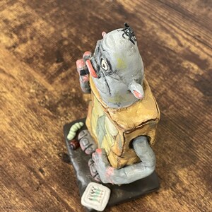 Puede incluir: Figura hecha a mano de una criatura con cabeza gris, vestida con un atuendo amarillo y verde. La figura tiene un peque&ntilde;o insecto en la cabeza y sostiene un objeto peque&ntilde;o. La base es negra y la figura tiene un estilo caprichoso y de arte popular.