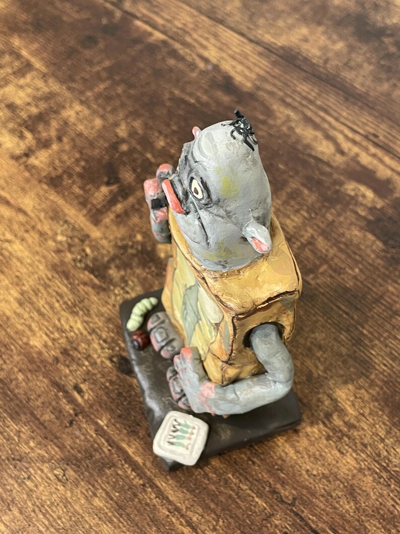 Puede incluir: Figura hecha a mano de una criatura con cabeza gris, vestida con un atuendo amarillo y verde. La figura tiene un peque&ntilde;o insecto en la cabeza y sostiene un objeto peque&ntilde;o. La base es negra y la figura tiene un estilo caprichoso y de arte popular.