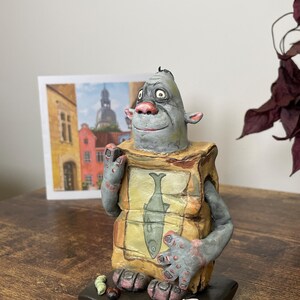 Puede incluir: Una escultura de cer&aacute;mica caprichosa de una criatura con cabeza gris, nariz rosa y brazos. Sostiene una caja con una ilustraci&oacute;n de pescado. La escultura se asienta sobre una base oscura con peque&ntilde;os detalles coloridos. Una acuarela est&aacute; en el fondo.