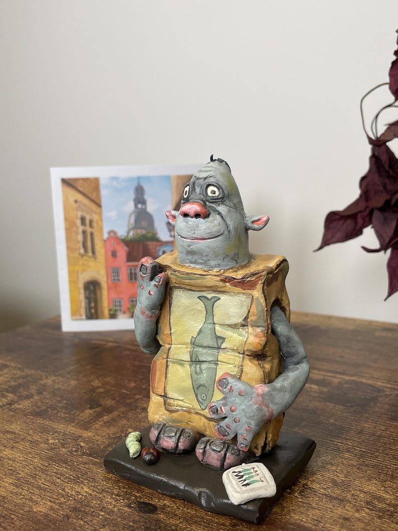 Puede incluir: Una escultura de cer&aacute;mica caprichosa de una criatura con cabeza gris, nariz rosa y brazos. Sostiene una caja con una ilustraci&oacute;n de pescado. La escultura se asienta sobre una base oscura con peque&ntilde;os detalles coloridos. Una acuarela est&aacute; en el fondo.