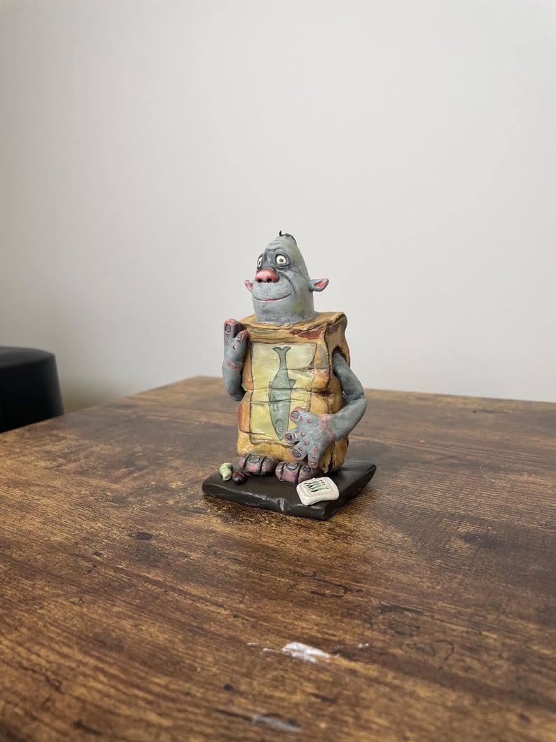 Puede incluir: Una figura caprichosa de una criatura gris con una gran nariz y orejas, que lleva un delantal con tem&aacute;tica de pescado. La figura se encuentra sobre una base negra con peque&ntilde;os detalles, incluyendo un peque&ntilde;o papel. El dise&ntilde;o es caricaturesco y detallado.