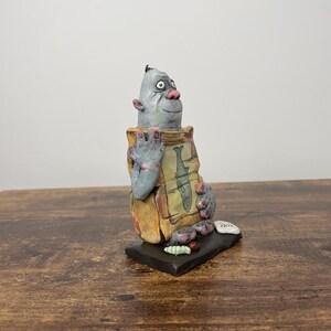 Puede incluir: Una escultura de cer&aacute;mica caprichosa de una criatura con cara gris, nariz rosa y orejas grandes. Sostiene una bolsa con ilustraciones de peces. La escultura est&aacute; sobre una base negra, con cuerpo marr&oacute;n y beige.