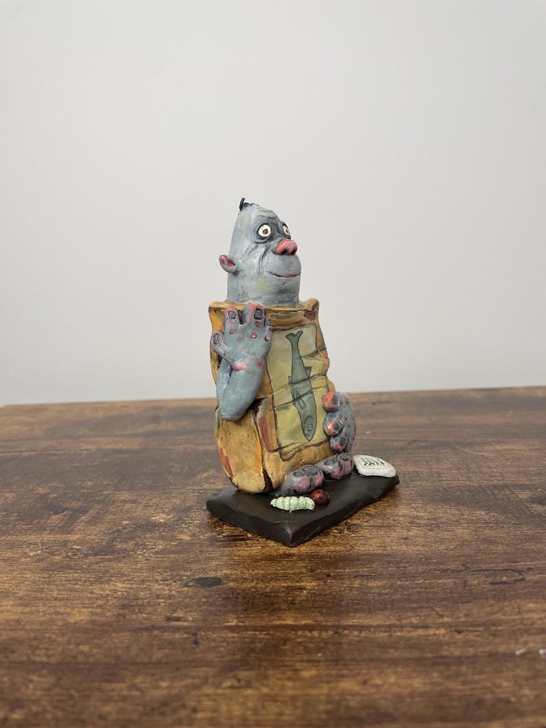 Puede incluir: Una escultura de cer&aacute;mica caprichosa de una criatura con cara gris, nariz rosa y orejas grandes. Sostiene una bolsa con ilustraciones de peces. La escultura est&aacute; sobre una base negra, con cuerpo marr&oacute;n y beige.