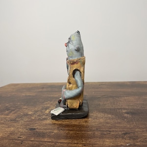 Puede incluir: Figura de cer&aacute;mica hecha a mano de una figura sentada con cabeza gris, nariz roja y gafas. La figura lleva un chaleco color canela y est&aacute; sentada sobre una base negra. Un peque&ntilde;o rect&aacute;ngulo blanco est&aacute; delante de la figura.