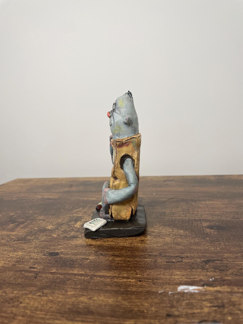 Puede incluir: Figura de cer&aacute;mica hecha a mano de una figura sentada con cabeza gris, nariz roja y gafas. La figura lleva un chaleco color canela y est&aacute; sentada sobre una base negra. Un peque&ntilde;o rect&aacute;ngulo blanco est&aacute; delante de la figura.