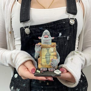 Puede incluir: Una escultura de cer&aacute;mica caprichosa de una criatura azul sosteniendo un paquete con una ilustraci&oacute;n de pescado. La escultura est&aacute; sobre una peque&ntilde;a base oscura con piezas tipo sushi y un peque&ntilde;o letrero. La criatura tiene ojos grandes y viste un atuendo amarillo y marr&oacute;n.