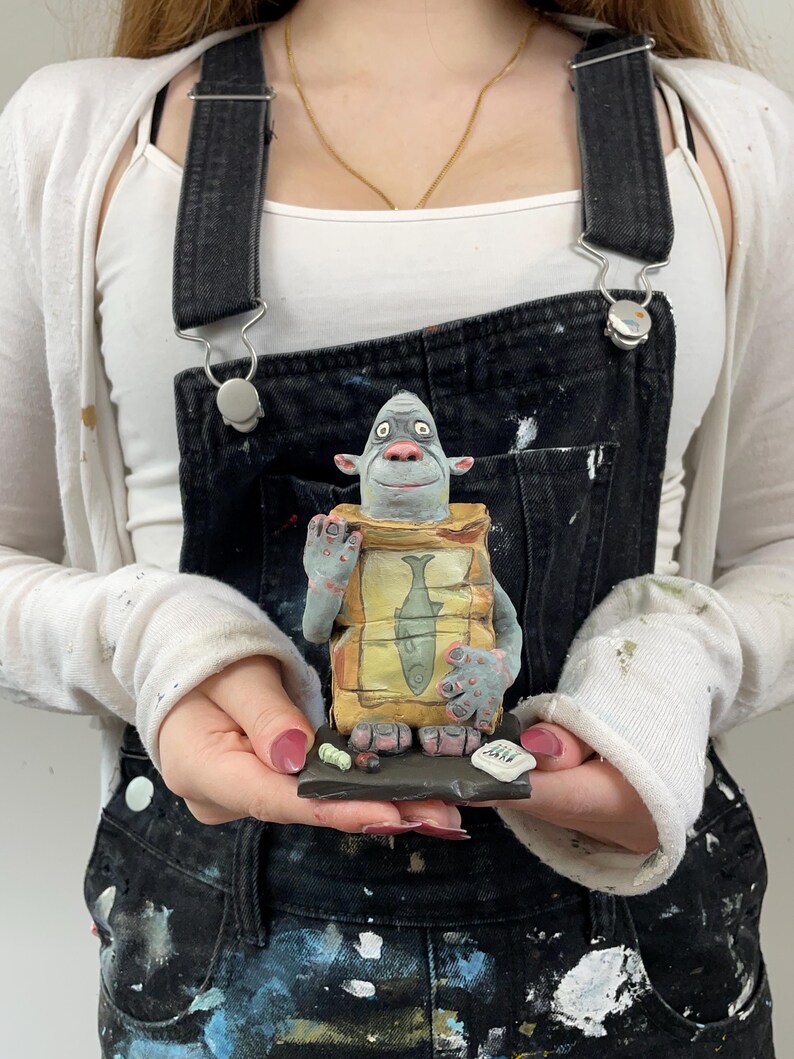 Puede incluir: Una escultura de cer&aacute;mica caprichosa de una criatura azul sosteniendo un paquete con una ilustraci&oacute;n de pescado. La escultura est&aacute; sobre una peque&ntilde;a base oscura con piezas tipo sushi y un peque&ntilde;o letrero. La criatura tiene ojos grandes y viste un atuendo amarillo y marr&oacute;n.