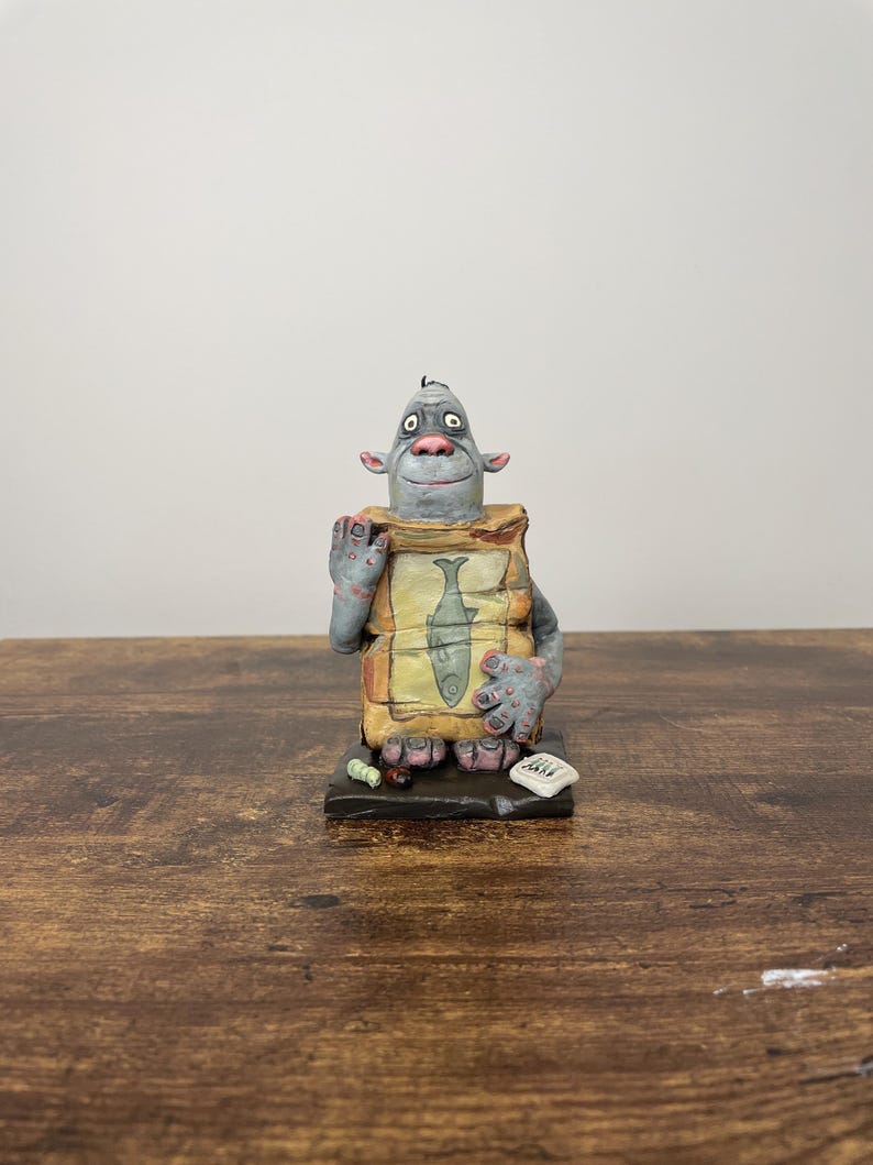 Puede incluir: Una figura caprichosa de una criatura gris con grandes orejas y una expresi&oacute;n amigable. Sostiene un letrero con una ilustraci&oacute;n de un pez. La base tiene peque&ntilde;os detalles coloridos. Una pieza de arte &uacute;nica, hecha a mano.