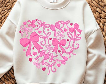 May include: A white sweatshirt featuring a heart design made of pink bows and small pink hearts. The bows vary in size and style, creating a playful and romantic aesthetic. The sweatshirt has a crew neck and long sleeves.