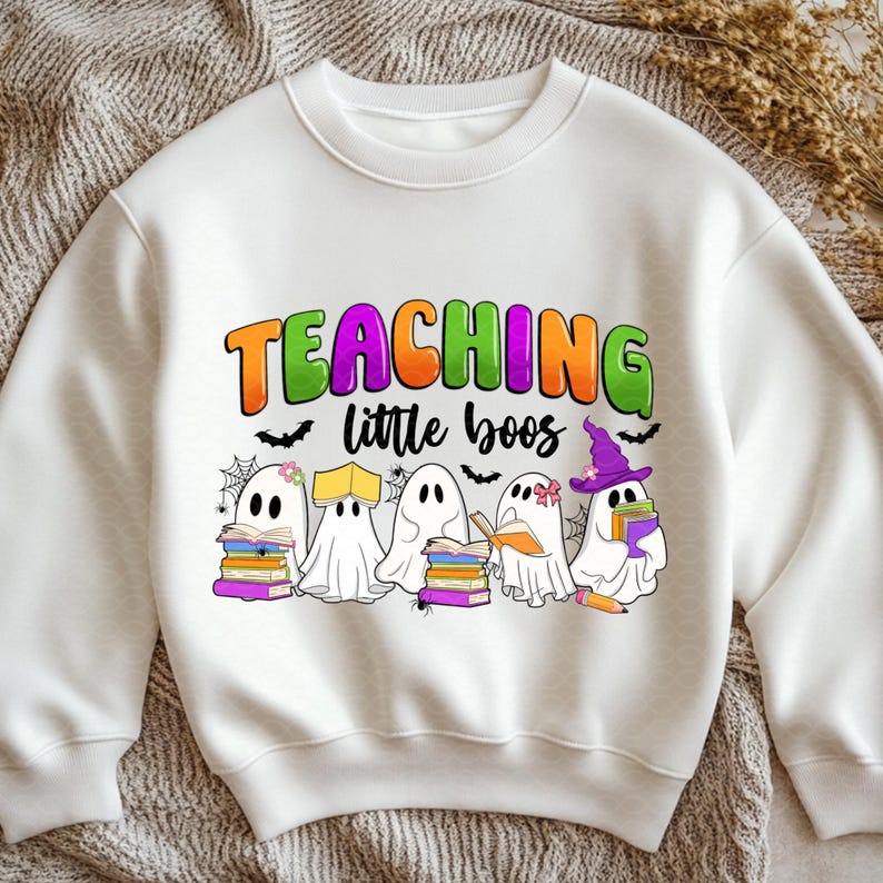 May include: White sweatshirt with a colorful graphic that reads "TEACHING little boos". The design features cartoon ghosts with books, spiderwebs, and bats, perfect for Halloween or a teacher's gift.