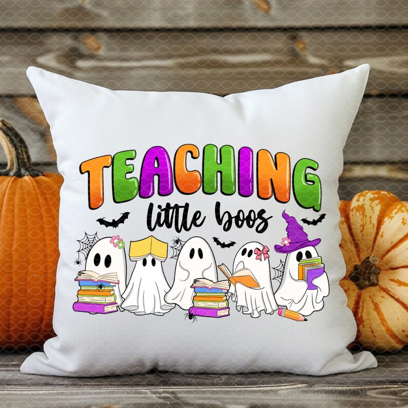 May include: White square pillow with the text "TEACHING little boos" in colorful letters. The pillow features cartoon ghosts reading books, with bats and spiderwebs. Pumpkins are in the background.