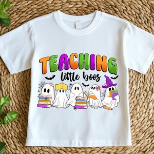 May include: White t-shirt featuring the text "TEACHING little boos" in orange, green, and purple. The design includes cartoon ghosts with books, bats, and spiderwebs, perfect for Halloween or a teacher.