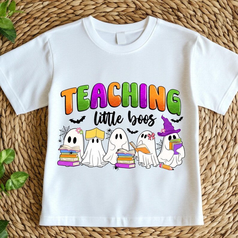 May include: White t-shirt featuring the text "TEACHING little boos" in orange, green, and purple. The design includes cartoon ghosts with books, bats, and spiderwebs, perfect for Halloween or a teacher.