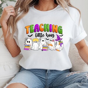 May include: White t-shirt with a colorful graphic that reads "TEACHING little boos." The design features cartoon ghosts holding books and wearing hats, with bats and spiderwebs. A fun Halloween-themed tee.