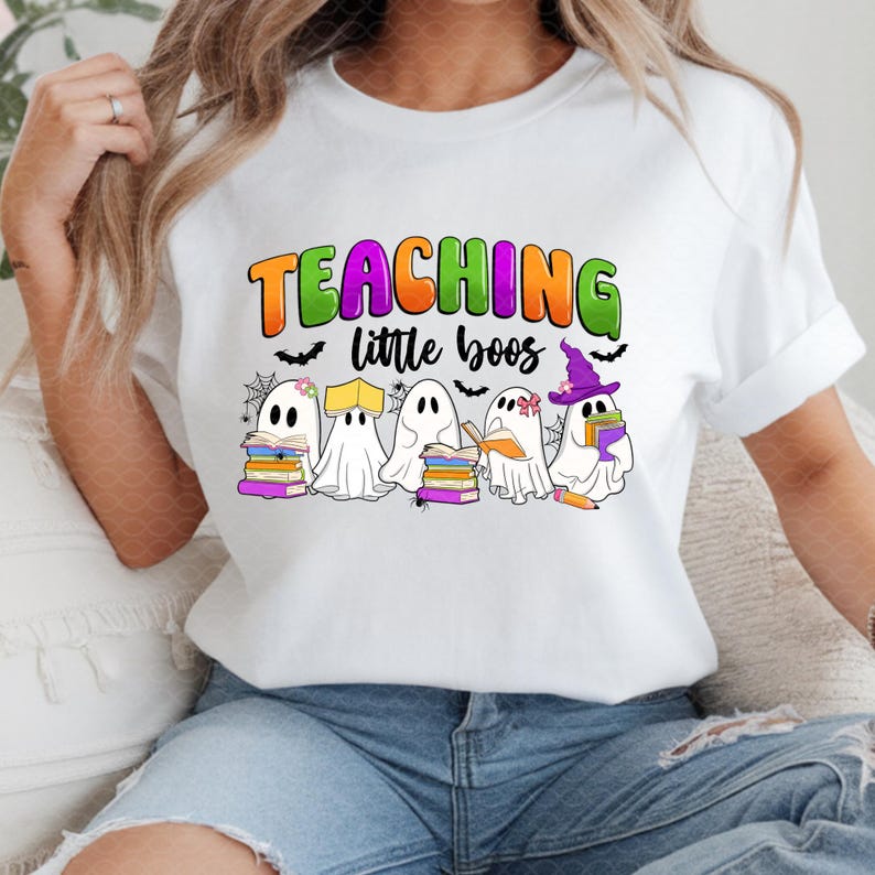 May include: White t-shirt with a colorful graphic that reads "TEACHING little boos." The design features cartoon ghosts holding books and wearing hats, with bats and spiderwebs. A fun Halloween-themed tee.