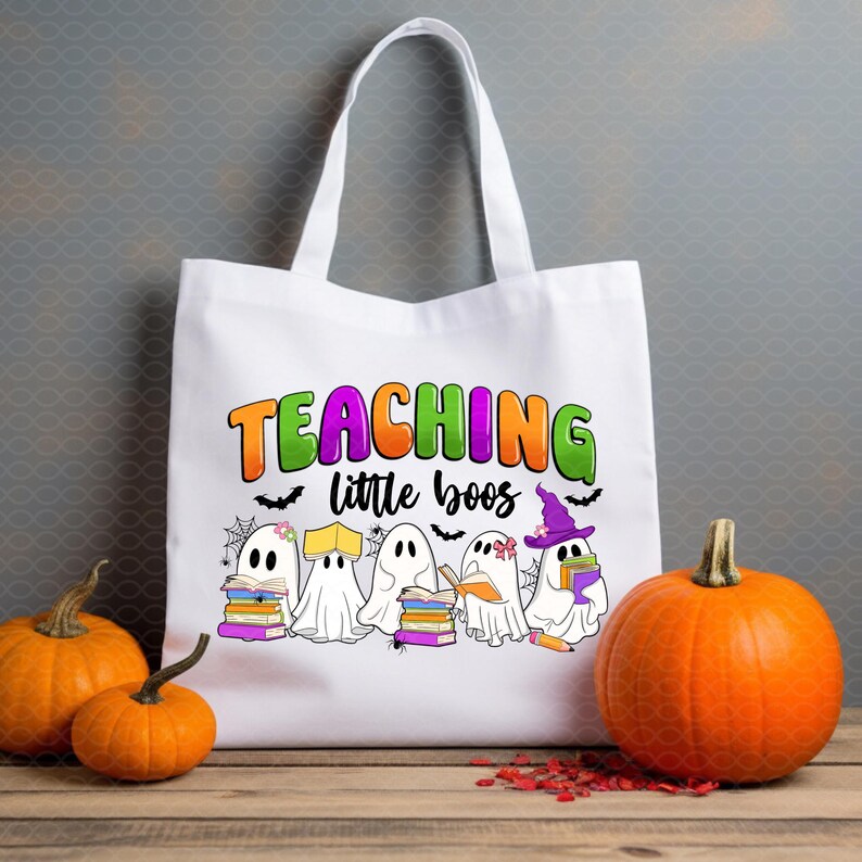 May include: White tote bag with the text "TEACHING little boos" in orange, green, and purple. The bag features cartoon ghosts holding books and wearing witch hats. Two orange pumpkins sit on a wooden surface.
