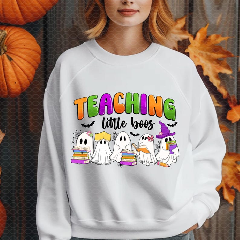 May include: White sweatshirt with the text "TEACHING little boos" in orange, green, and purple. The design features cartoon ghosts holding books and wearing witch hats, perfect for Halloween or teachers.