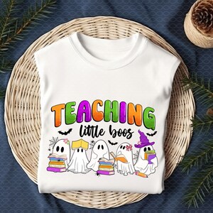 May include: White t-shirt featuring the text "TEACHING little boos" in orange, green, and purple. The design includes cartoon ghosts with books, bats, and spiderwebs, perfect for Halloween or a teacher's gift.