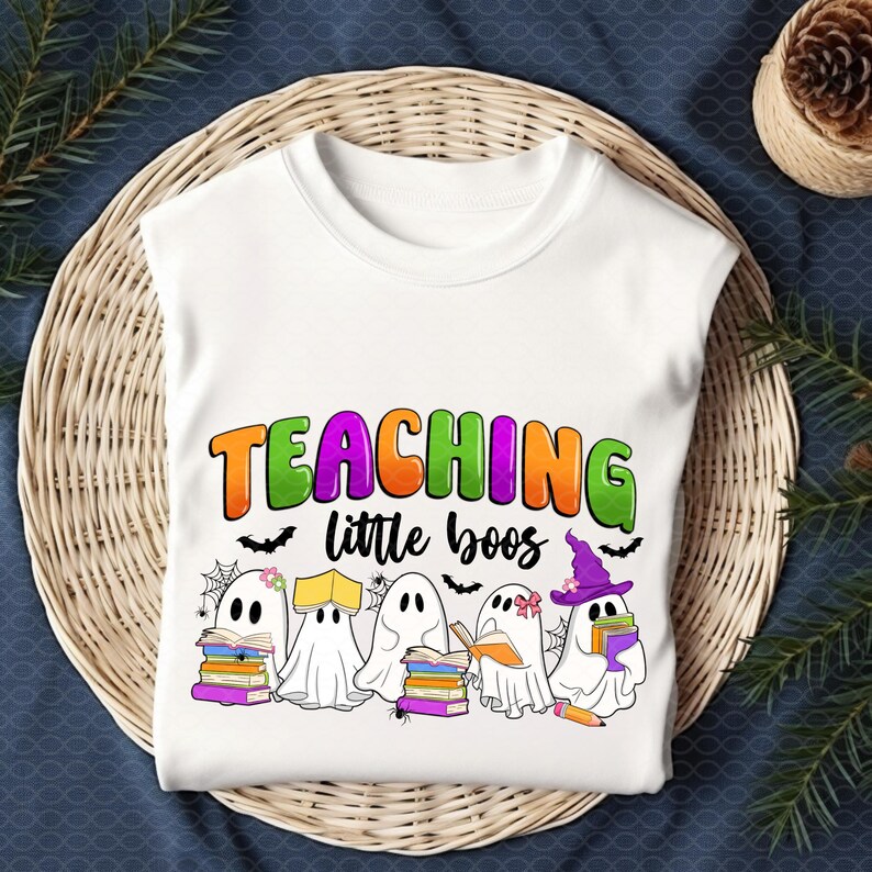 May include: White t-shirt featuring the text "TEACHING little boos" in orange, green, and purple. The design includes cartoon ghosts with books, bats, and spiderwebs, perfect for Halloween or a teacher's gift.