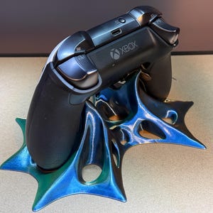 May include: A black Xbox controller rests on a blue and green 3D-printed stand. The stand has a unique, spiky design with a gradient of colors. The controller is black with the Xbox logo.