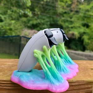 Op de afbeelding: Een witte gamecontroller rust op een aangepaste standaard. De standaard heeft een kleurrijk ontwerp met een basis van roze, blauw en groen, met groene ranken die de controller ondersteunen. De controller heeft zwarte knoppen.