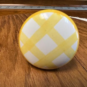 May include: A round, white and yellow cabinet knob with a plaid pattern. The knob has a glossy finish and is set against a wooden surface. The yellow lines create a grid-like design.