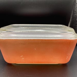 May include: A rectangular orange glass storage container with a clear glass lid. The container features a solid matte orange finish on the exterior sides and a smooth, transparent glass cover that sits flush on top of the rectangular base.