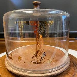 May include: A clear glass cake dome with a wooden base and a wooden knob on top. The glass dome has gold decorative accents. The cake stand is sitting on a wooden surface.