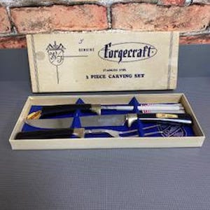 May include: A vintage Forgecraft 3-piece carving set in its original box. The set includes a carving knife, a carving fork, and a sharpening steel. The box is beige with blue text and a logo. The set is displayed on a blue surface.
