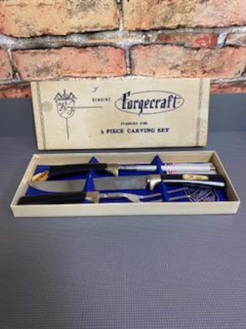 May include: A vintage Forgecraft 3-piece carving set in its original box. The set includes a carving knife, a carving fork, and a sharpening steel. The box is beige with blue text and a logo. The set is displayed on a blue surface.