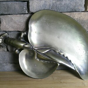 May include: A metallic brass-toned serving tray shaped like a lobster with two distinct concave sections. The lobster figure is integrated into the design, forming the handle and central divider of the tray, featuring a smooth, polished finish.