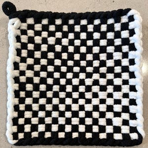 Puede incluir: Un salvamanteles cuadrado tejido con un patrón de tablero de ajedrez en blanco y negro. Los bordes están ribeteados con un borde blanco en forma de cuerda, y un lazo negro está unido a la esquina superior para colgarlo. El salvamanteles mide unos 20 cm cuadrados.