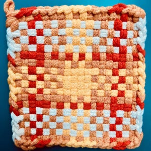 May include: Woven square hot pad with a checkerboard design. The colours are red, orange, tan, and white. It has a braided edge and a hanging loop. The hot pad is made from a thick, durable material, suitable for kitchen use.