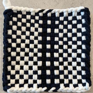 May include: A square, woven potholder in a checkerboard pattern of navy blue and white. The edges are trimmed with a navy blue and white braided border. A small white loop is attached to one corner for hanging.