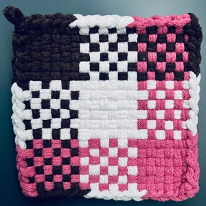 May include: Square, woven hot pad in brown, white, and pink. The design features a checkerboard pattern in the center, with a brown border and a loop for hanging. The hot pad is made of thick, durable material.